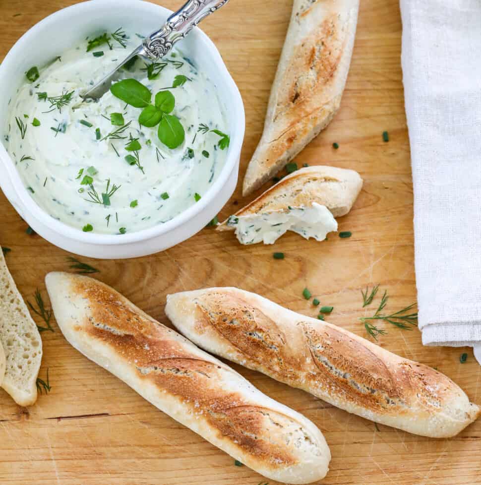 A wooden cutting board with small baguettes sliced open with a white bowl of whipped creamed cheese spread on a slice.