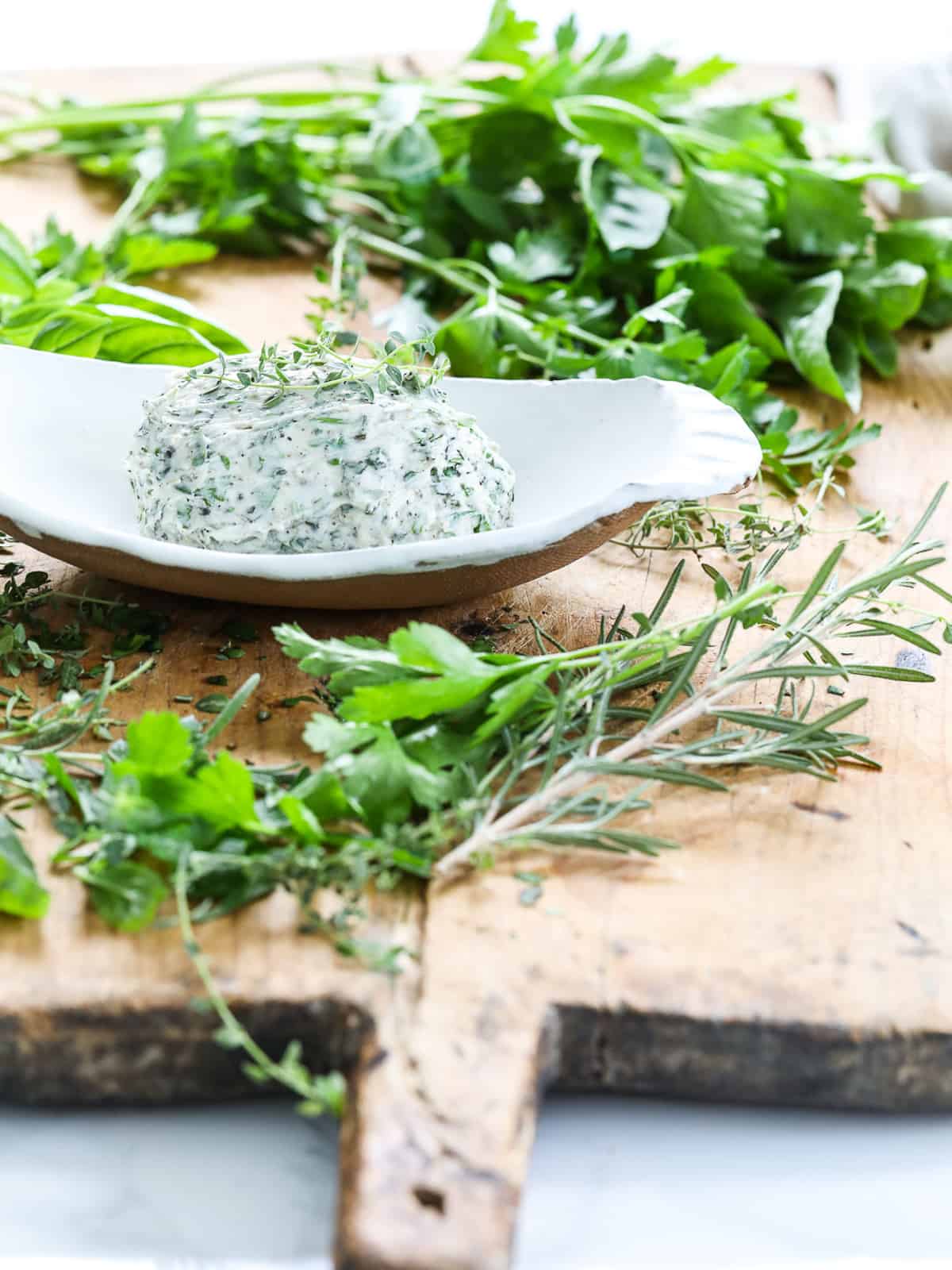 A white ceramic dish homemade herb butter and fresh herbs on a cutting board.