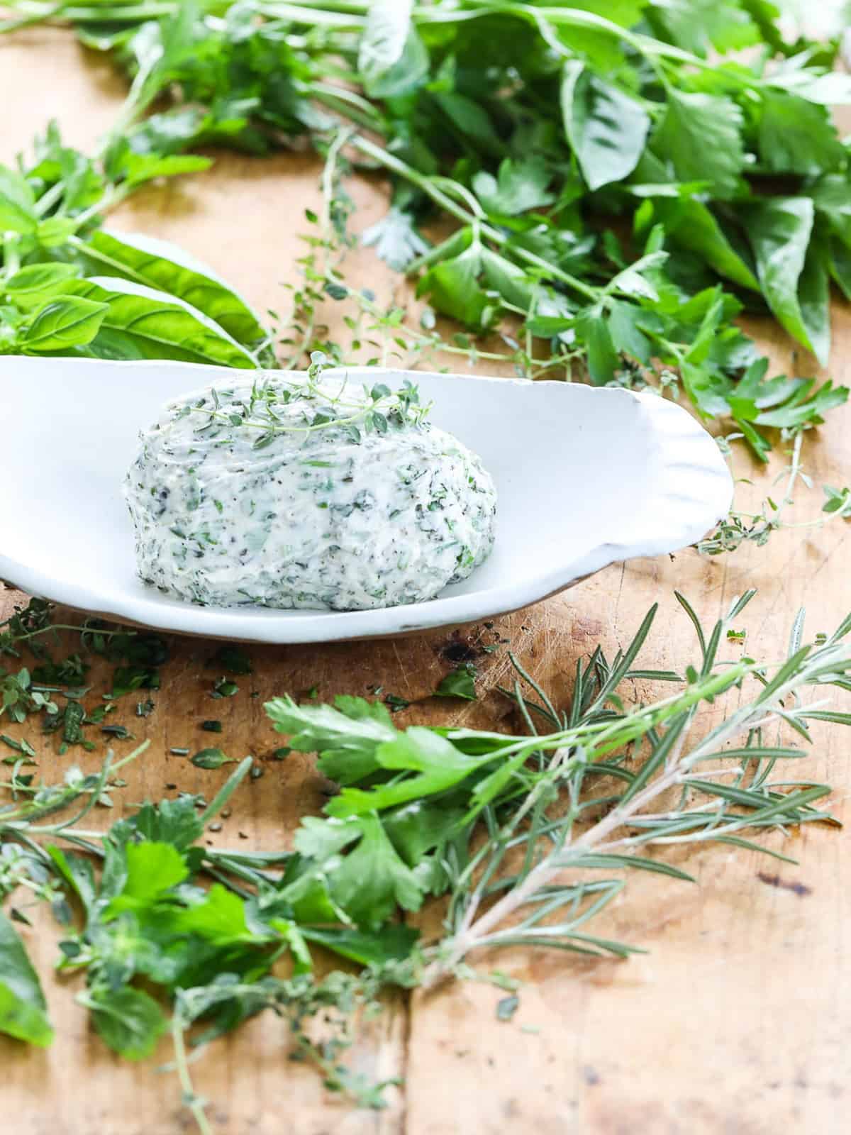 Fresh herb butter in a white dish on a cutting board with herbs.