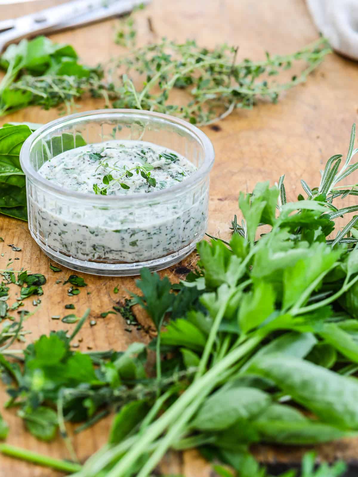 Herb butter in a small glass container on a cutting board with herbs.