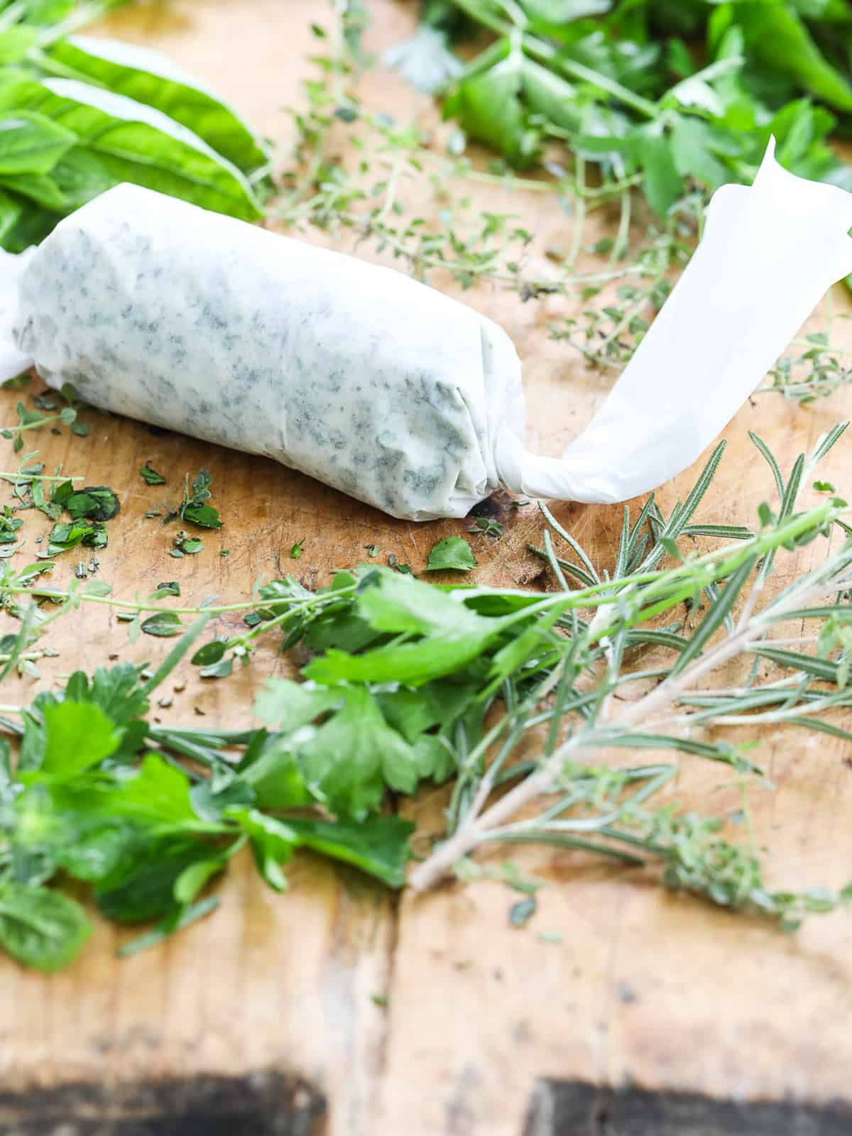 Herb butter wrapped in a piece of white parchment paper on a cutting board with fresh herbs.