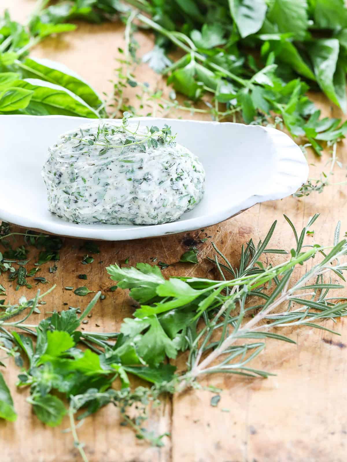Fresh herb butter in a white dish on a cutting board with herbs.