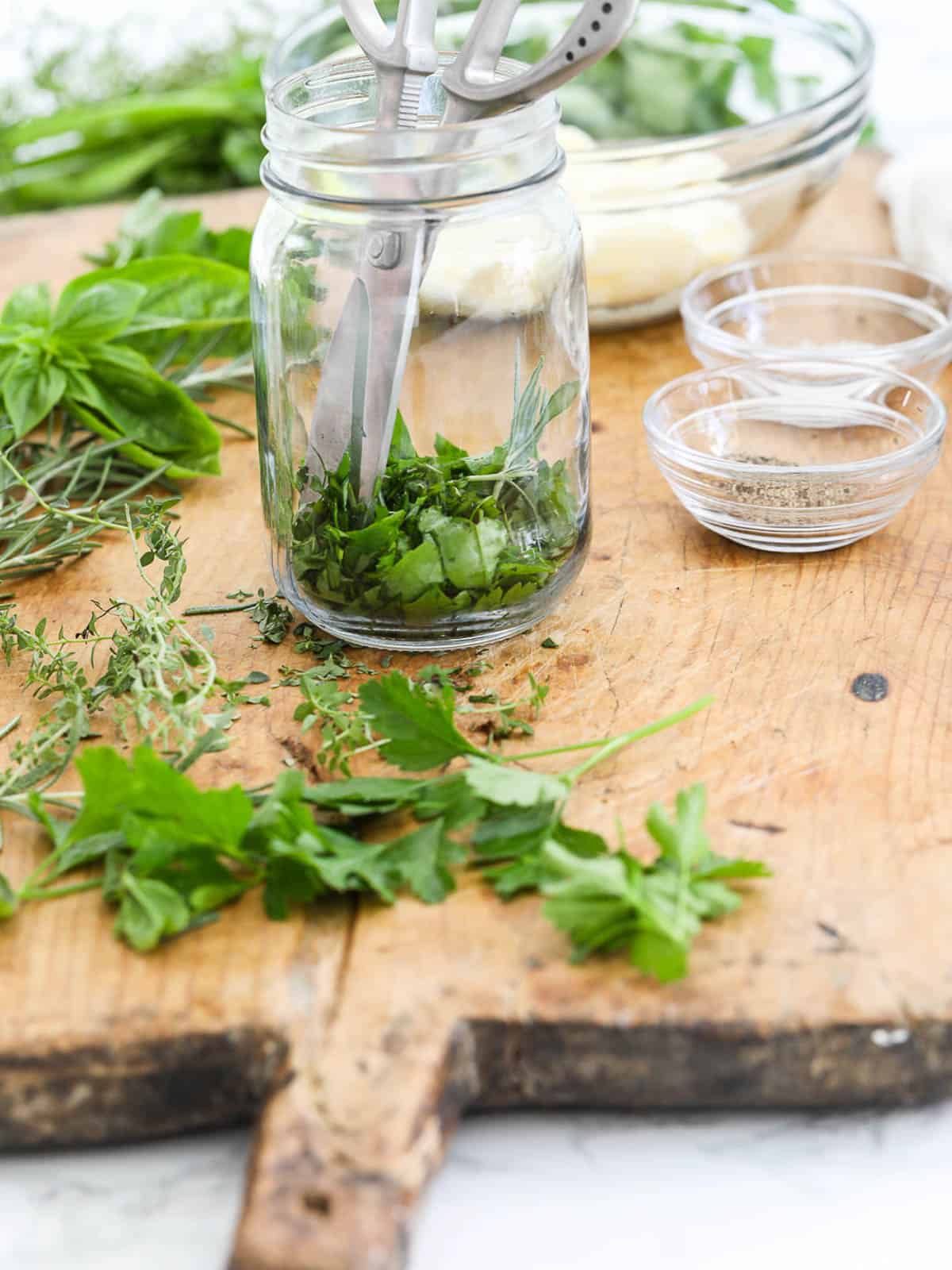 A pair of silver kitchen scissors in a glass jar with fresh herbs and butter to making herb butter.