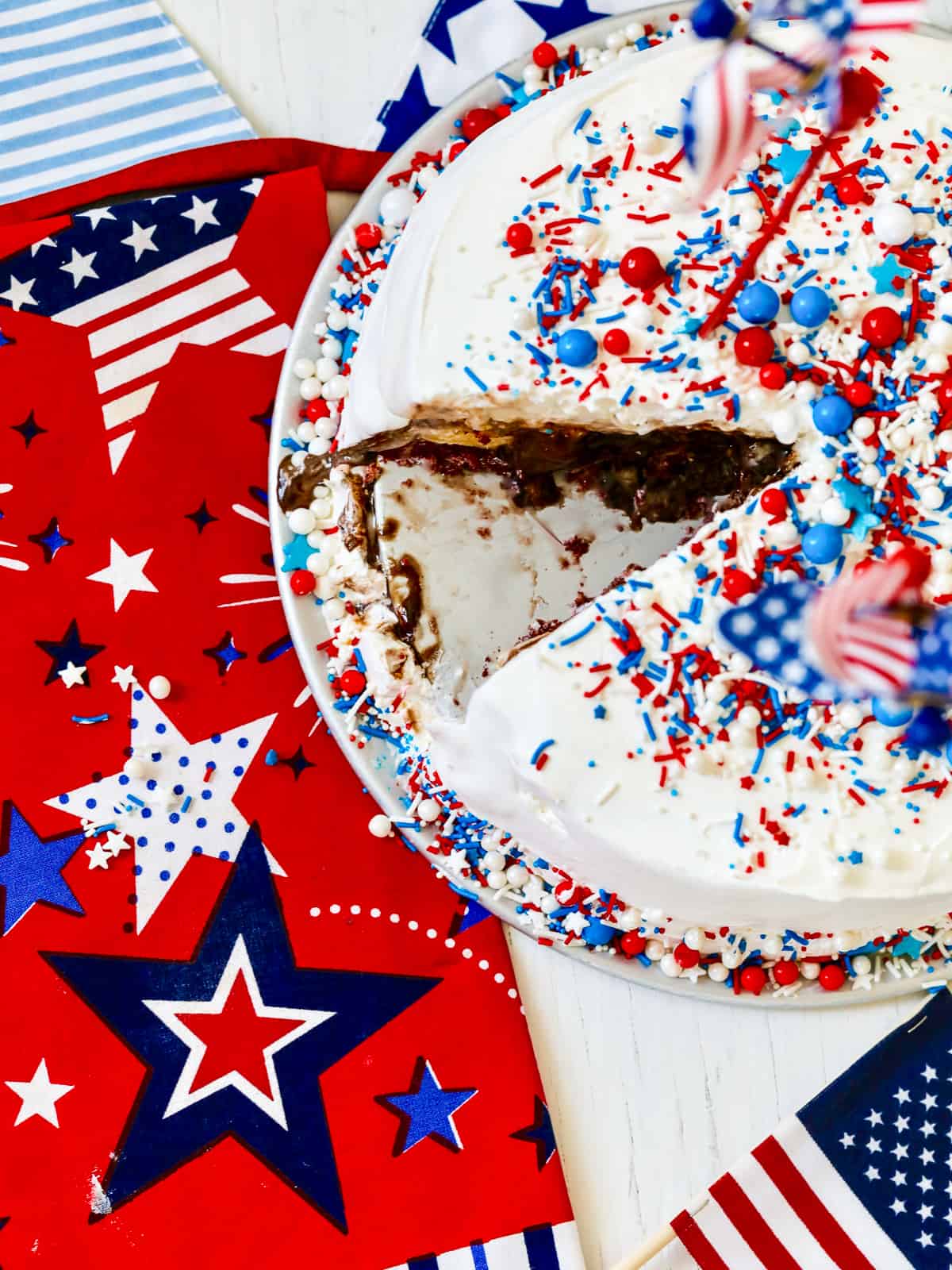 An ice cream cake for July 4th with a slice cut out of it on a patriotic red, white, and blue star tablecloth.