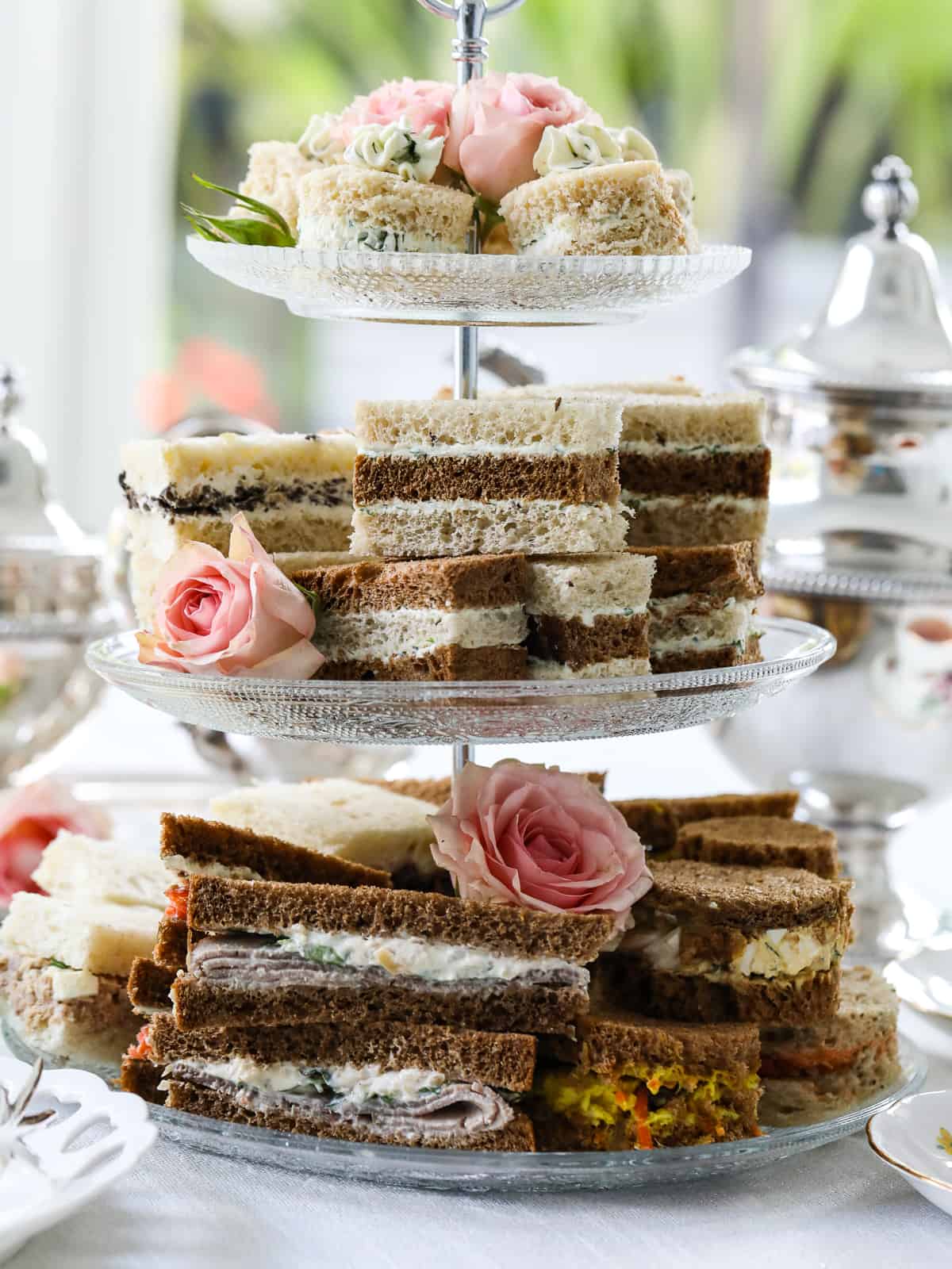 Side view of a triple glass tiered tray with stacks of tea sandwiches sliced in different shapes and colors of white and dark bread.
