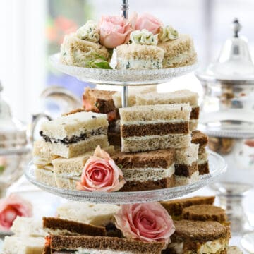 Tea sandwiches stacked on a 3 tiered glass tray at a tea party with assorted fillings and flavors.