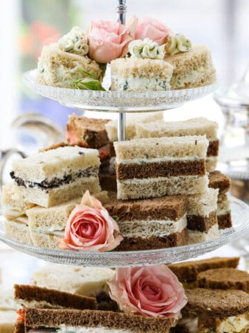 Tea sandwiches stacked on a 3 tiered glass tray at a tea party with assorted fillings and flavors.