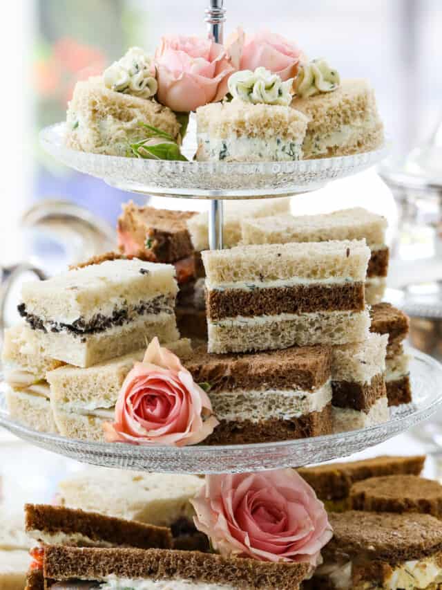 Tea sandwiches stacked on a 3 tiered glass tray at a tea party with assorted fillings and flavors.