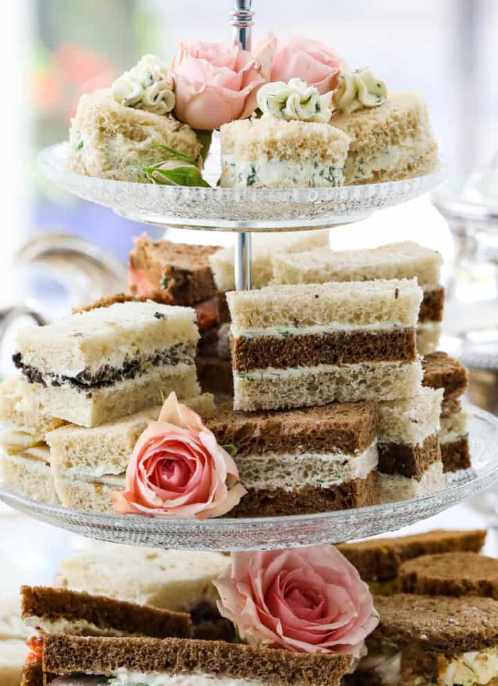 Tea sandwiches stacked on a 3 tiered glass tray at a tea party with assorted fillings and flavors.