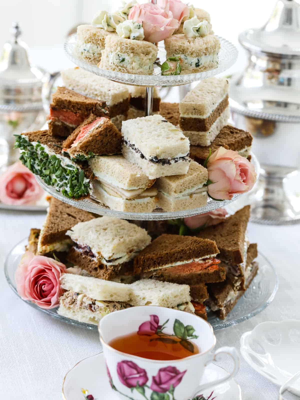 An assortment of different tea sandwiches on a tiered glass tray with different fillings.