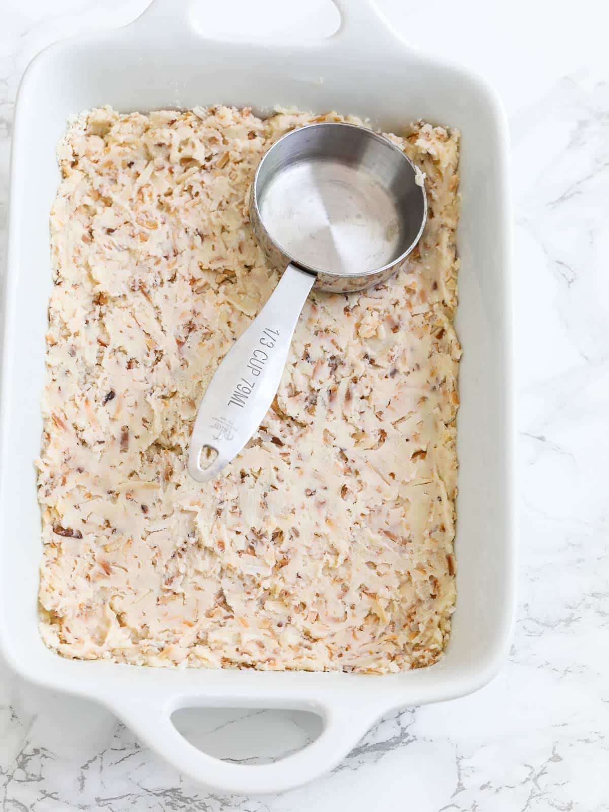 A measuring cup pressing the coconut cookie crust into the pan for lemon bars. 