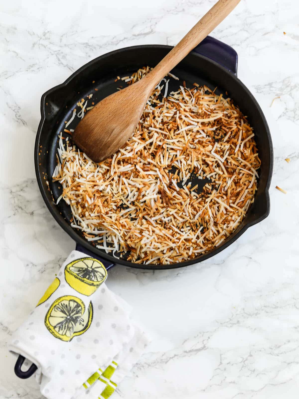 Toasted coconut in a cast iron skillet with a lemon decorated towel around the handle and a wooden spoon. 