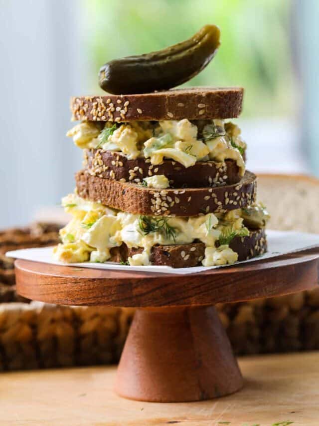 Egg salad made into an egg sandwich on brown bread stacked on a wood stand with a pickle on top.