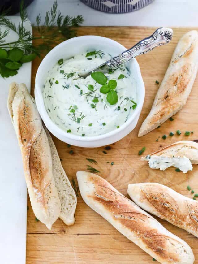 A wood cutting board with a white ceramic bowl of herb cream cheese dip and small baguettes.