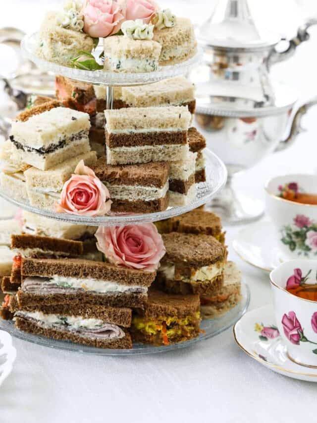 A 3 tiered glass tea tray stacked with small sandwiches with two tea cups.