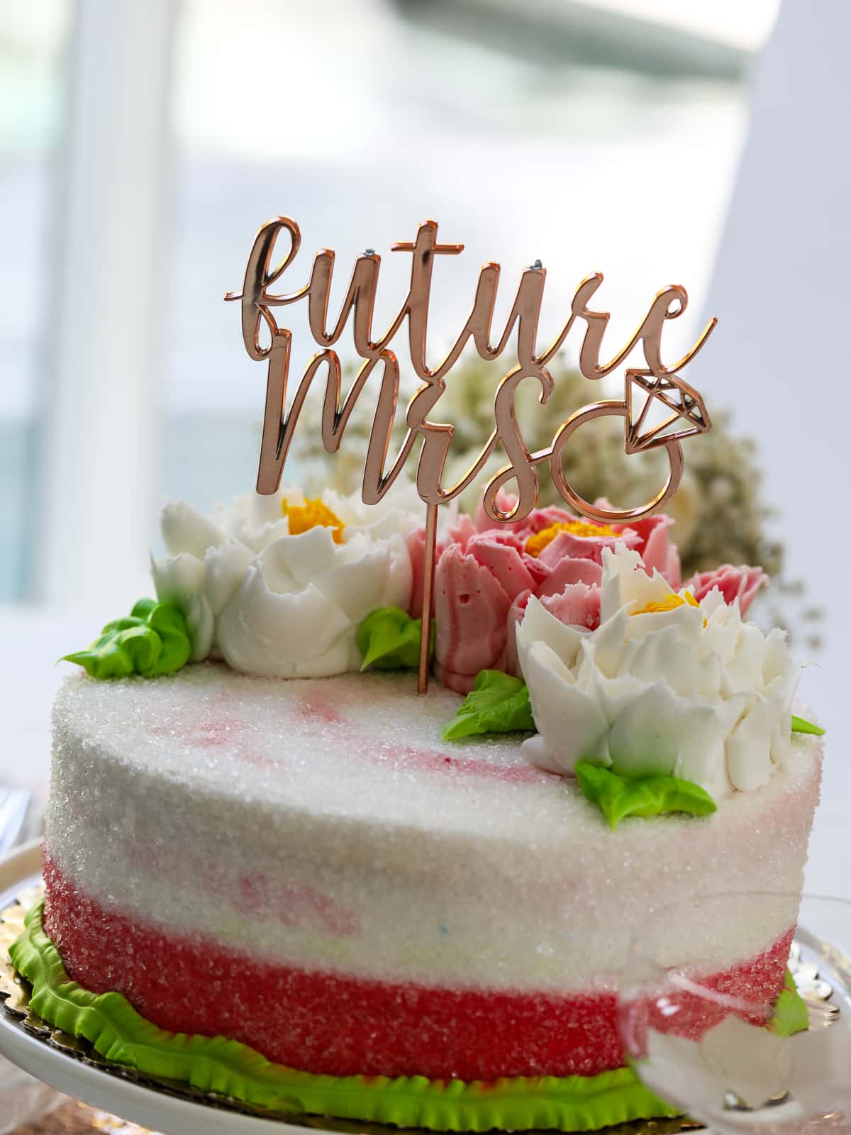 Close up of a white cake topped with pastel frosing flowers and a decoration that says "future Mrs" at a bridal shower party