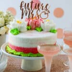 A pink and white cake with buttercream flowers on a cake plate with glasses of pink champagne at a bridal shower.