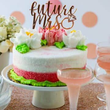 A pink and white cake with buttercream flowers on a cake plate with glasses of pink champagne at a bridal shower.