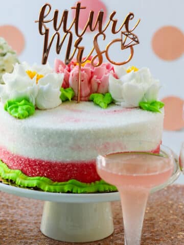 A pink and white cake with buttercream flowers on a cake plate with glasses of pink champagne at a bridal shower.