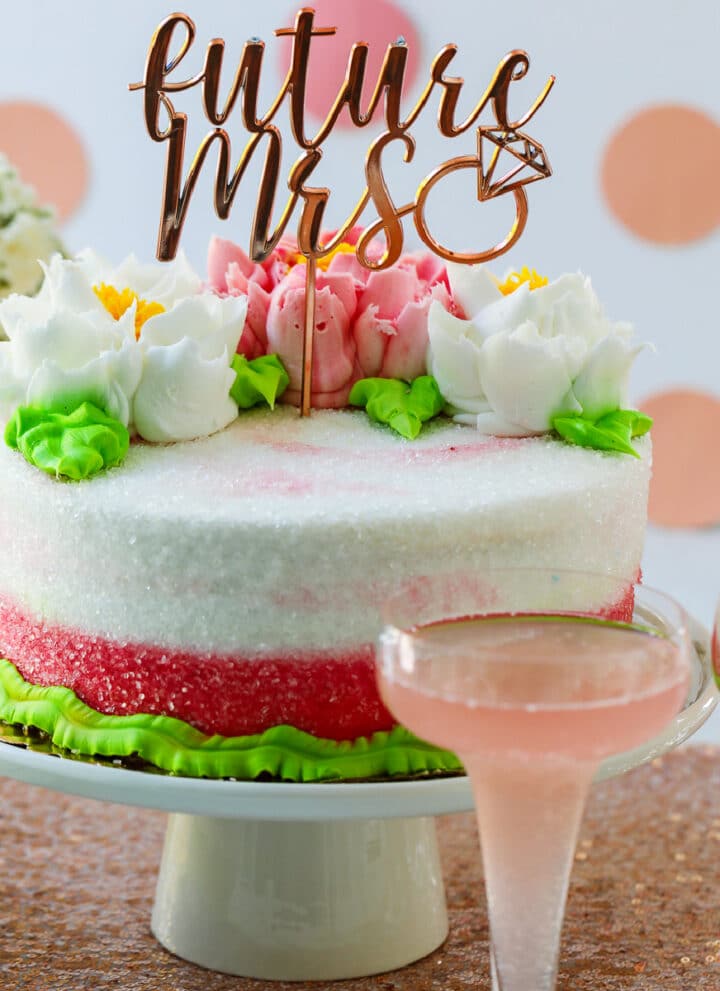 A pink and white cake with buttercream flowers on a cake plate with glasses of pink champagne at a bridal shower.