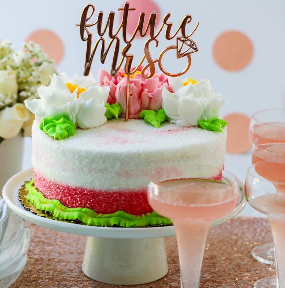 A pink and white cake with buttercream flowers on a cake plate with glasses of pink champagne at a bridal shower.