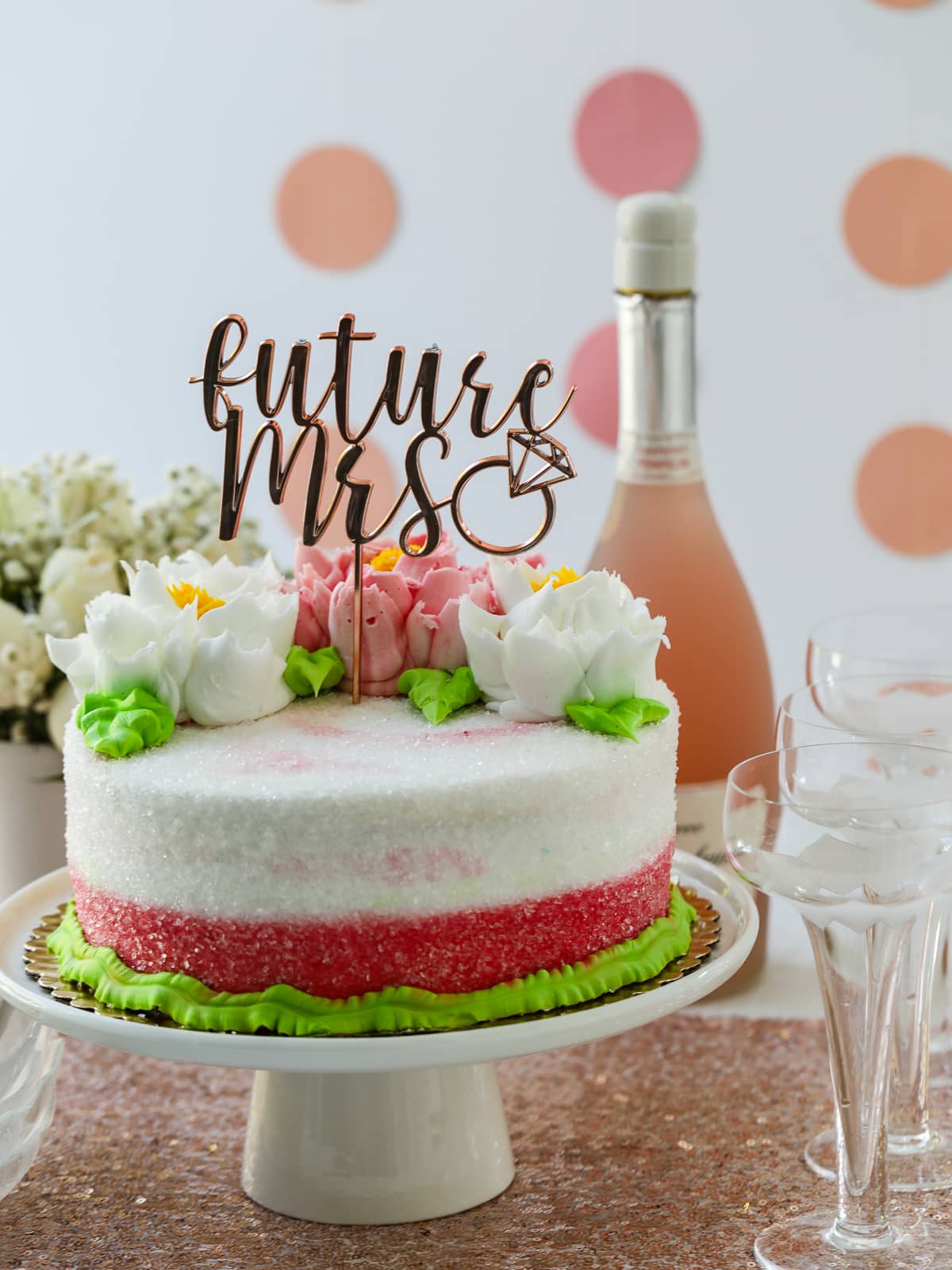 A white cake topped with a "future Mrs" decoration and a bottle of pink champagne with glasses at a bridal shower party. 