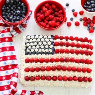 An American flag cake decorated with blueberries, raspberries, and white frosting with a patriotic star towel.