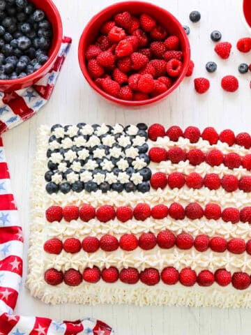 An American flag cake decorated with blueberries, raspberries, and white frosting with a patriotic star towel.