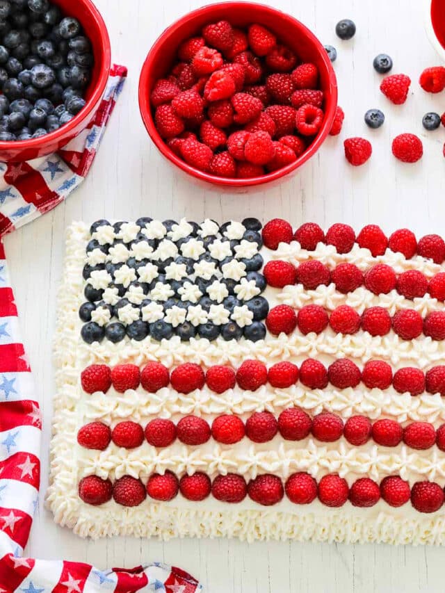 An American flag cake decorated with blueberries, raspberries, and white frosting with a patriotic star towel.