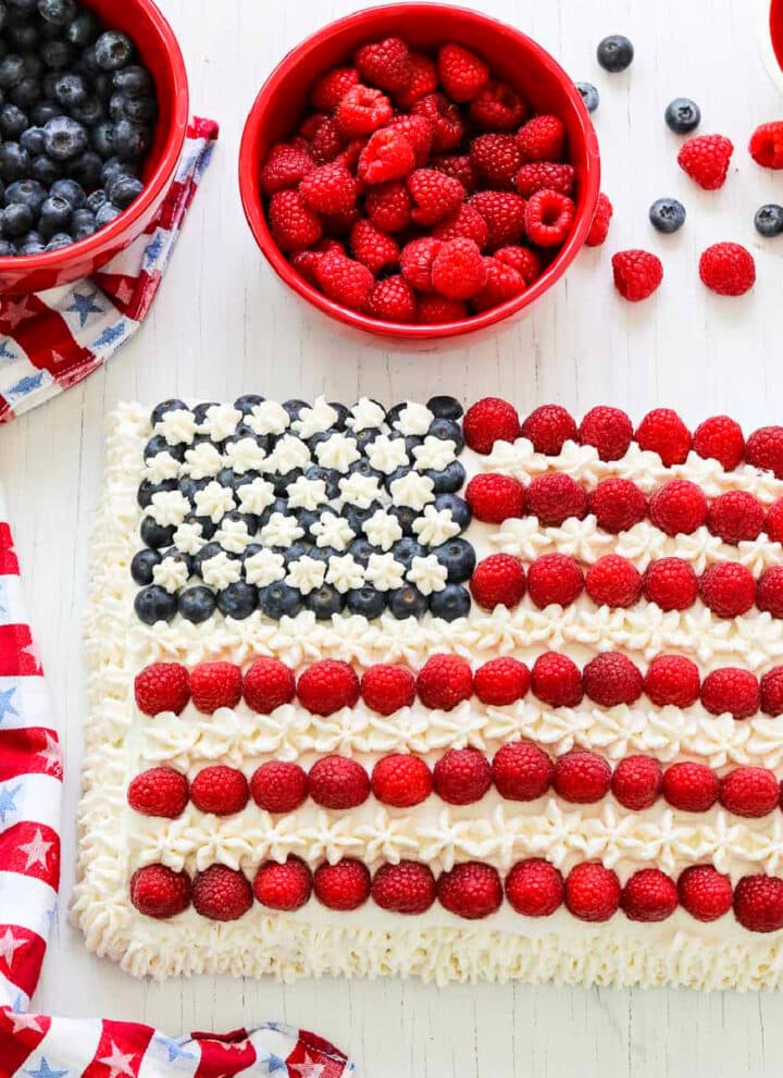 An American flag cake decorated with blueberries, raspberries, and white frosting with a patriotic star towel.