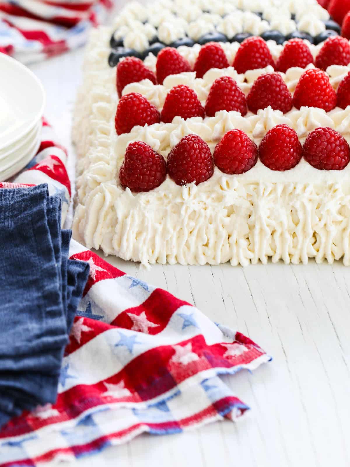 The corner of a white frosted cake decorated with raspberries with a patriotic towel. 