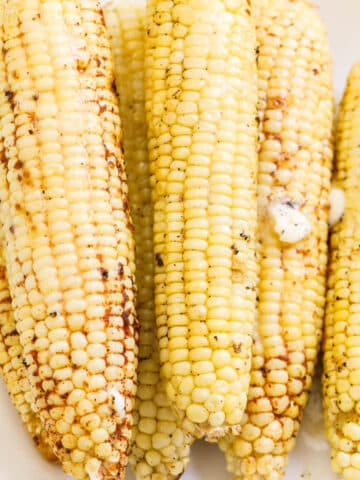 Ears of grilled corn on a white platter seasoned with melted butter on top.