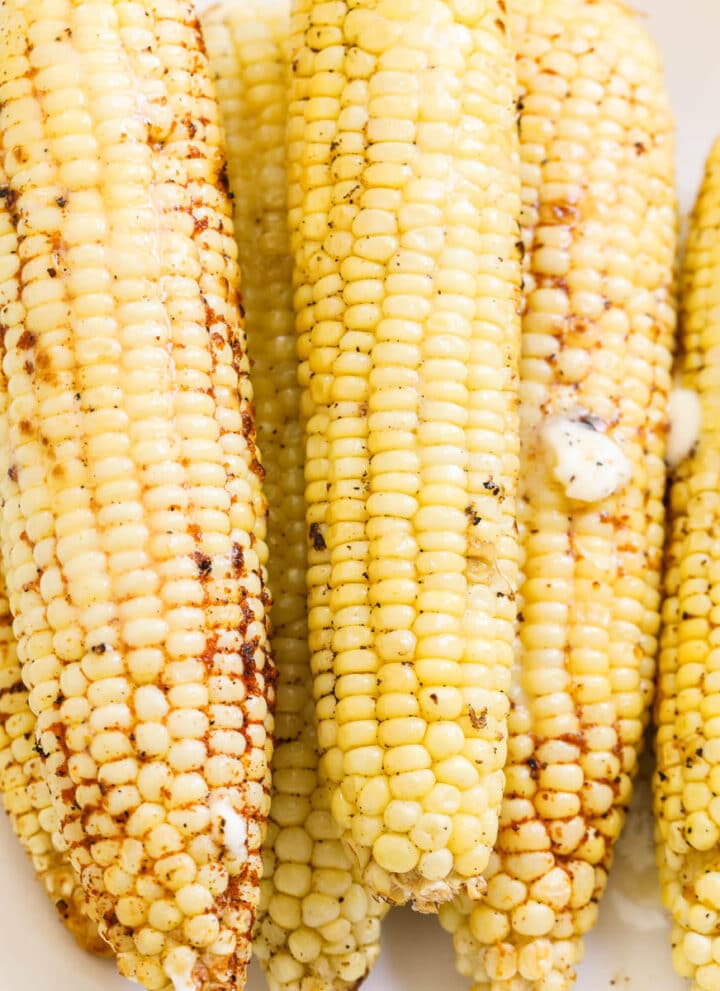 Ears of grilled corn on a white platter seasoned with melted butter on top.