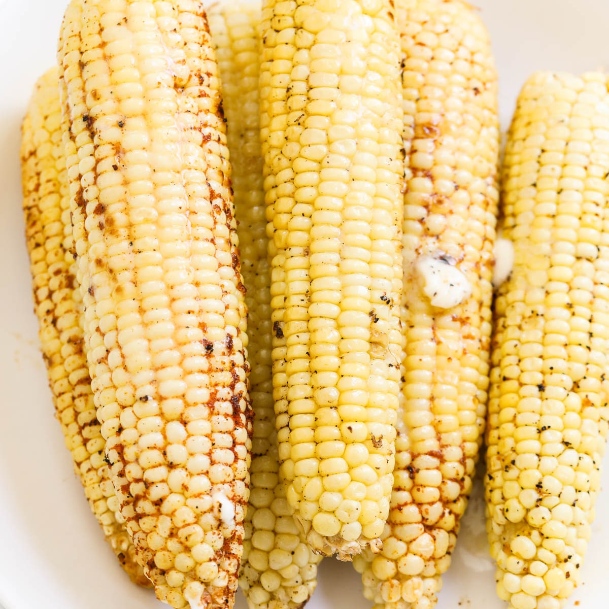 Ears of grilled corn on a white platter seasoned with melted butter on top.