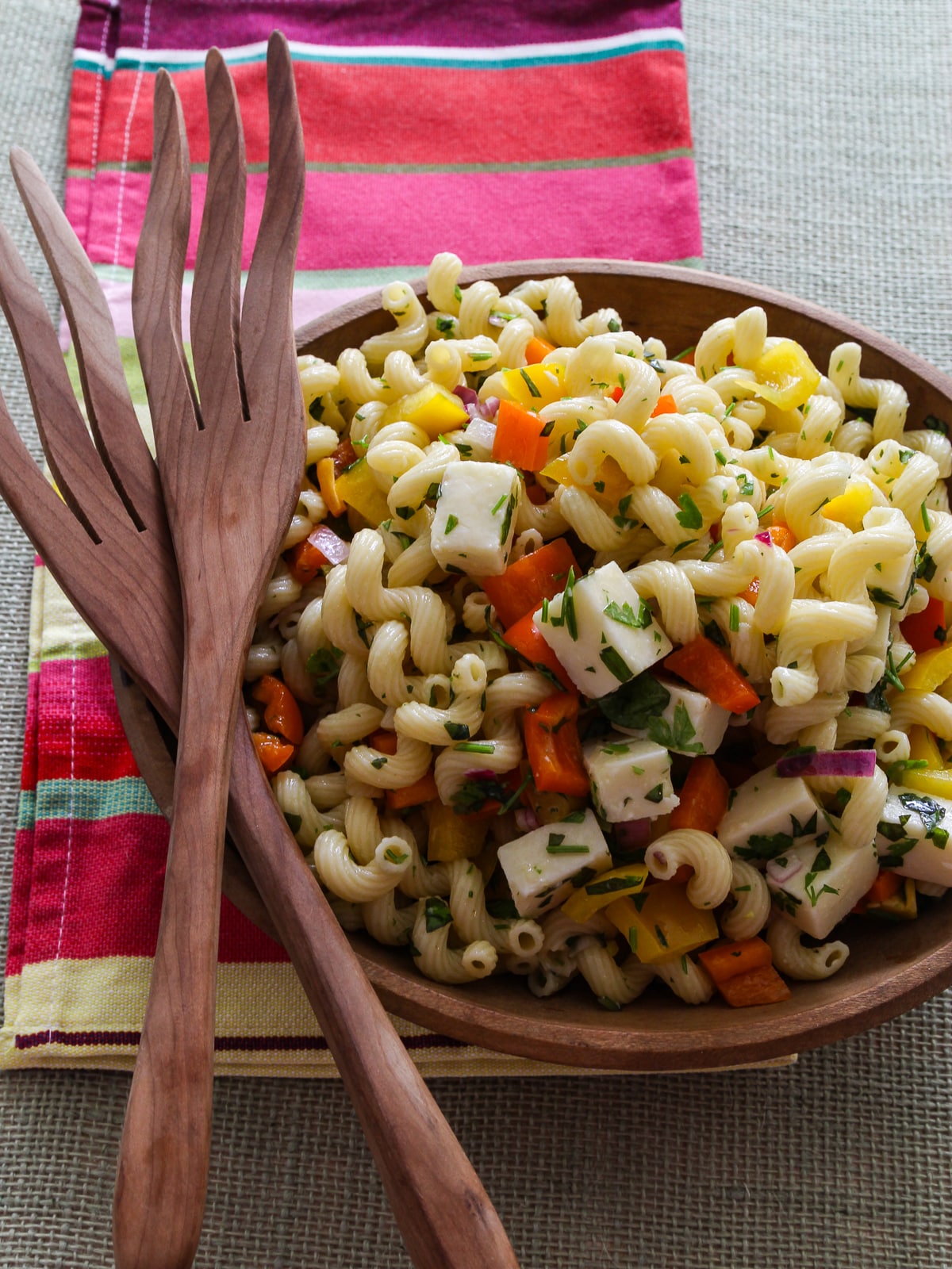 Pasta salad in a wood bowl with serving wood tongs on a colorful towel. 