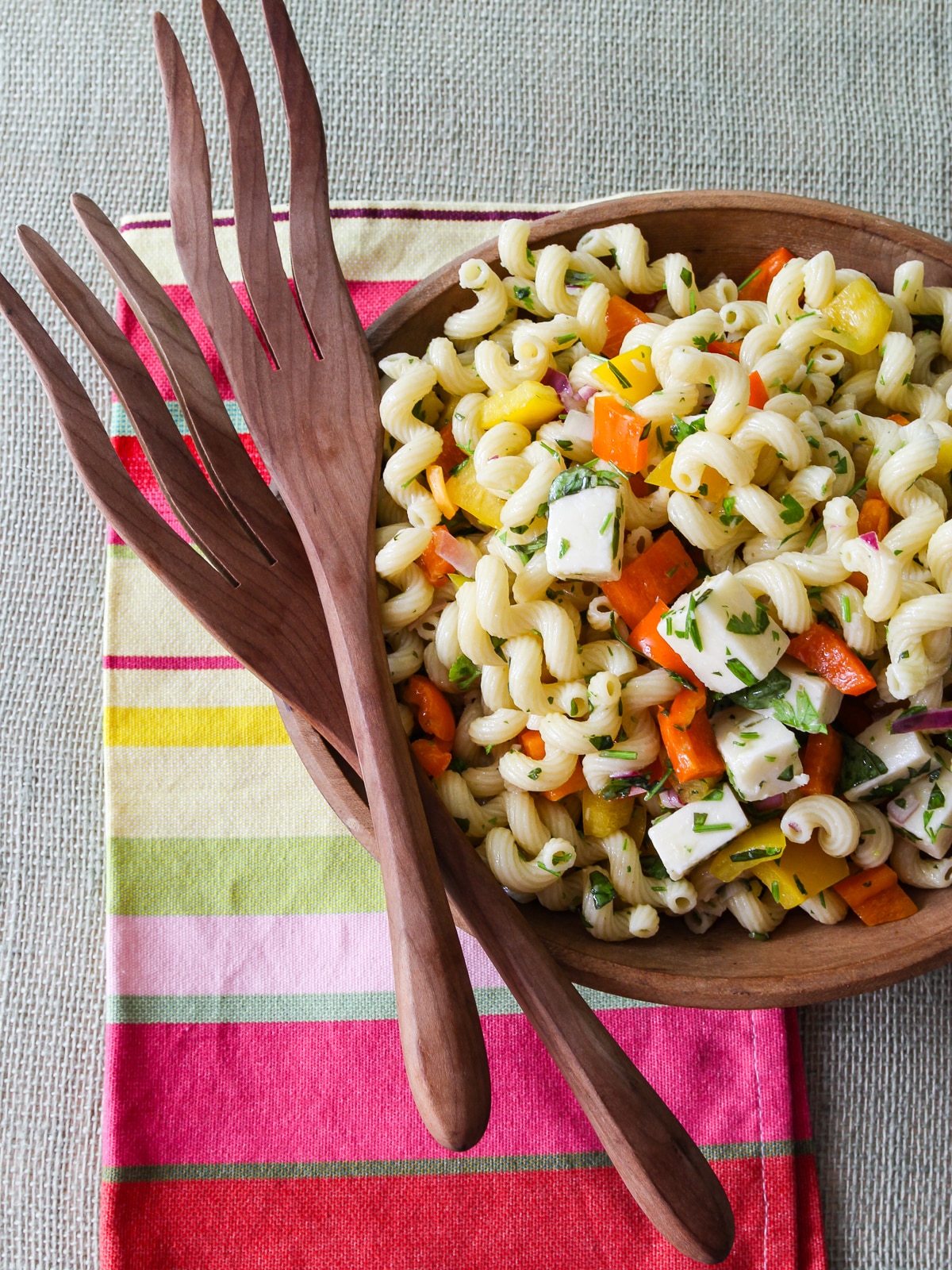 A wood bowl with pasta salad and wood tongs resting on the edge on a colorful striped towel. 