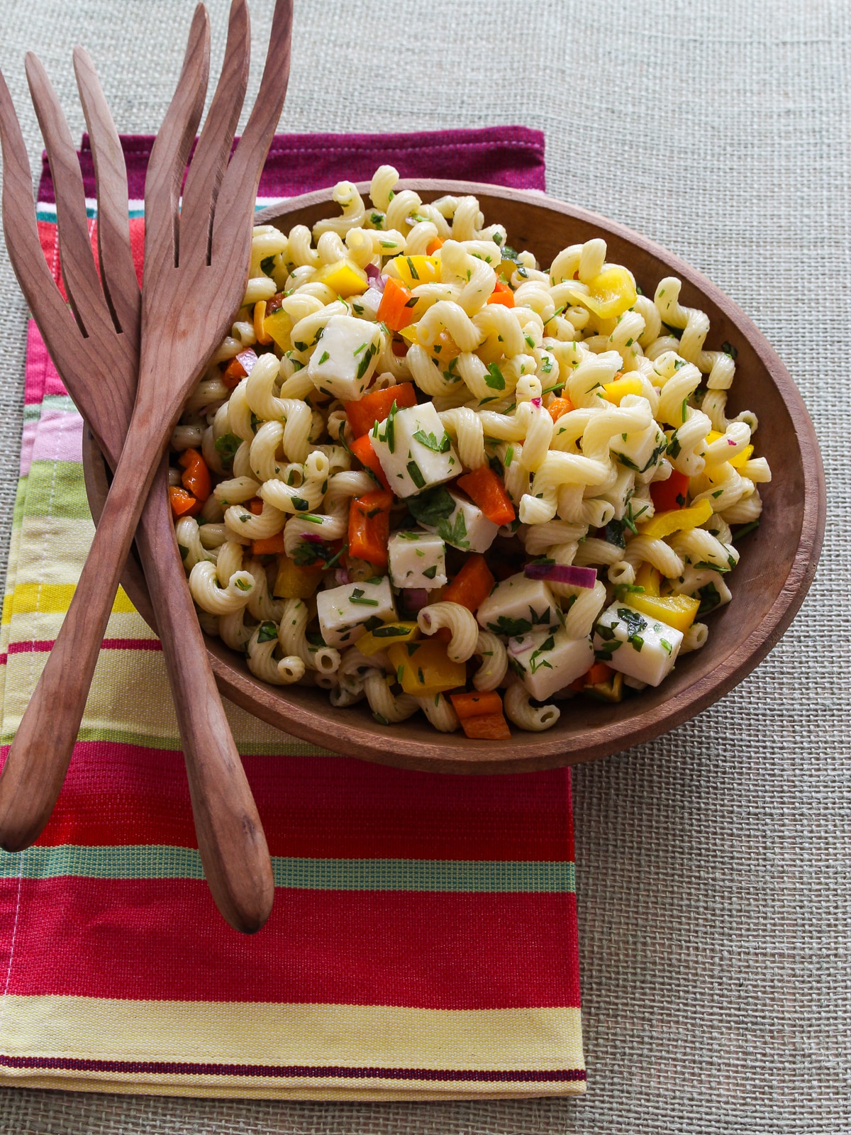 A wood bowl and tongs with pasta salad recipe inside on a colorful striped towel. 