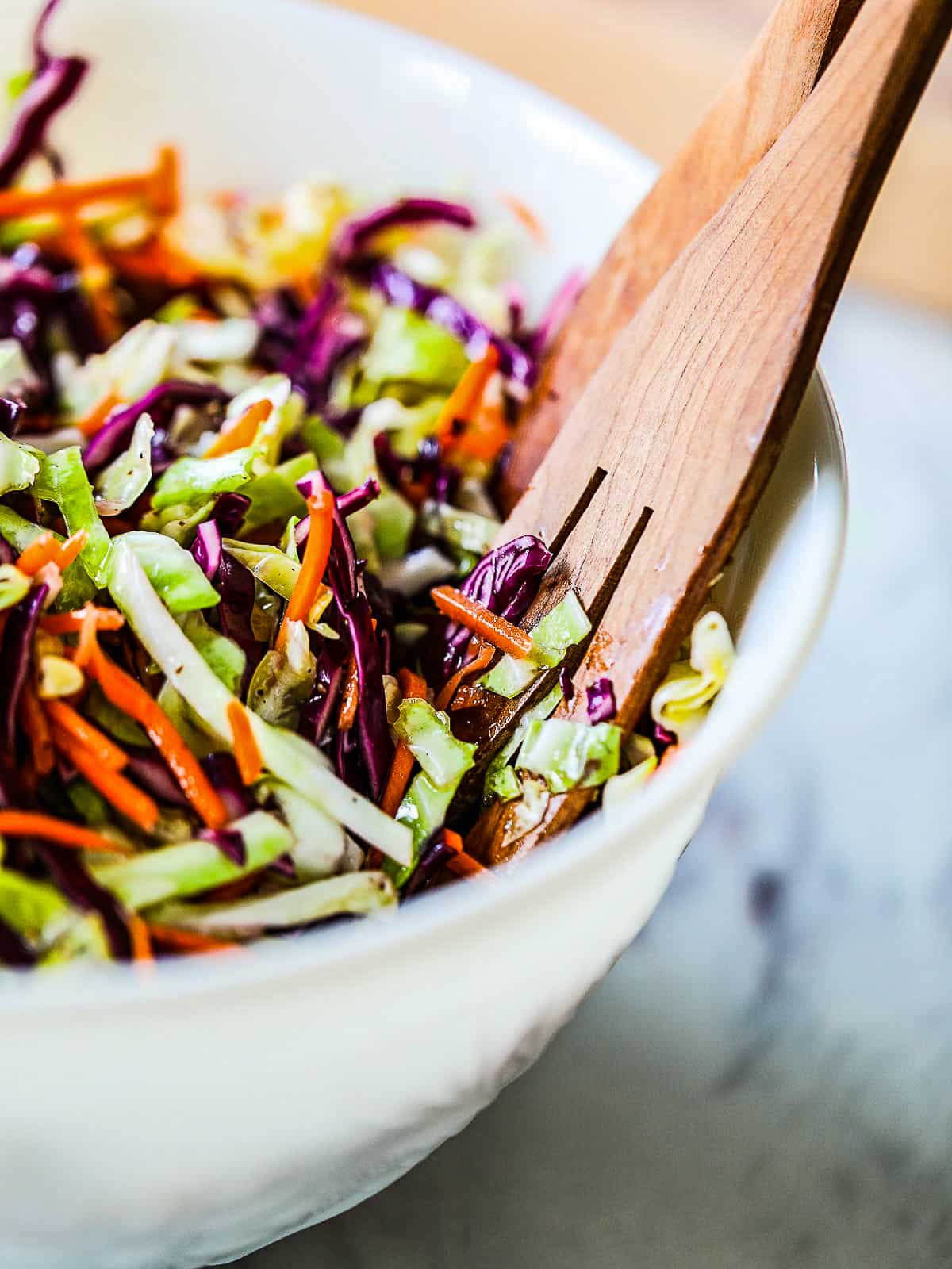 A white bowl filled with colorful tri color cole slaw with salad tongs.