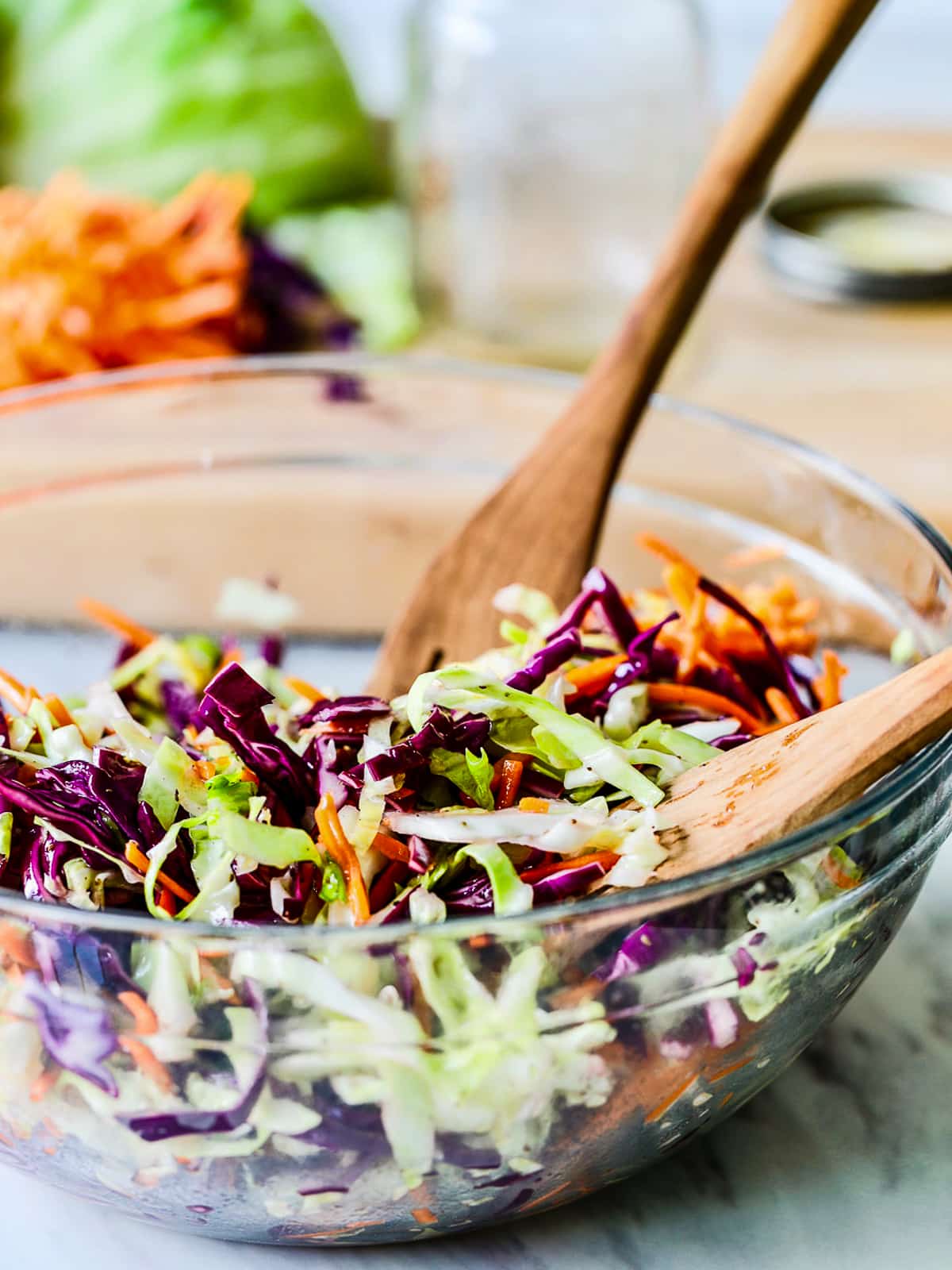 Coleslaw in a clear bowl tossed with wood serving tongs in a kitchen. 