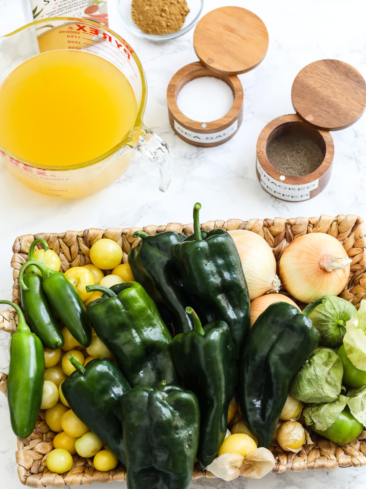 The ingredients to make green enchilada sauce on a table with spices, chicken stock, and peppers in a basket. 