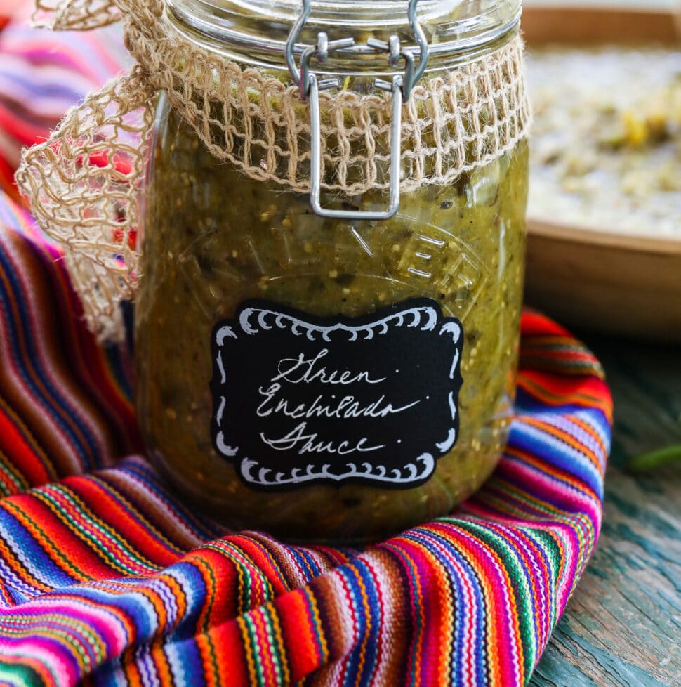A glass jar with a clamp filled with green enchilada sauce on a colorful striped piece of cloth.