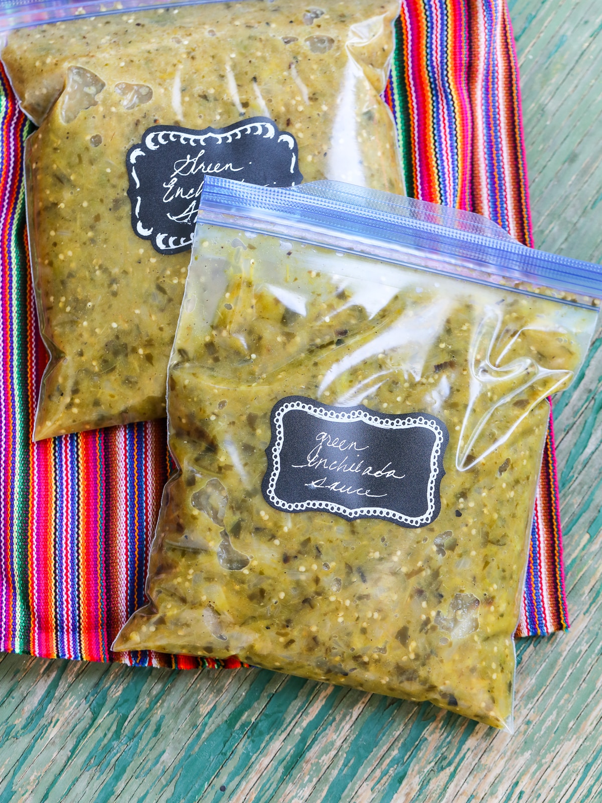 Two zip topped plastic freezer bags filled with green enchilada sauce on a colorful striped piece of cloth.