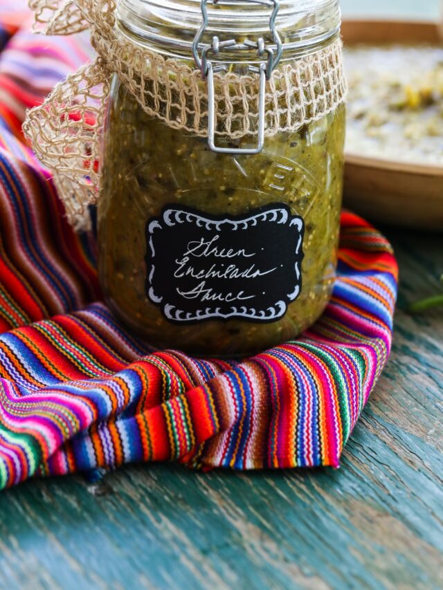 A clear glass jar filled with green enchilada sauce on a colorful striped cloth.
