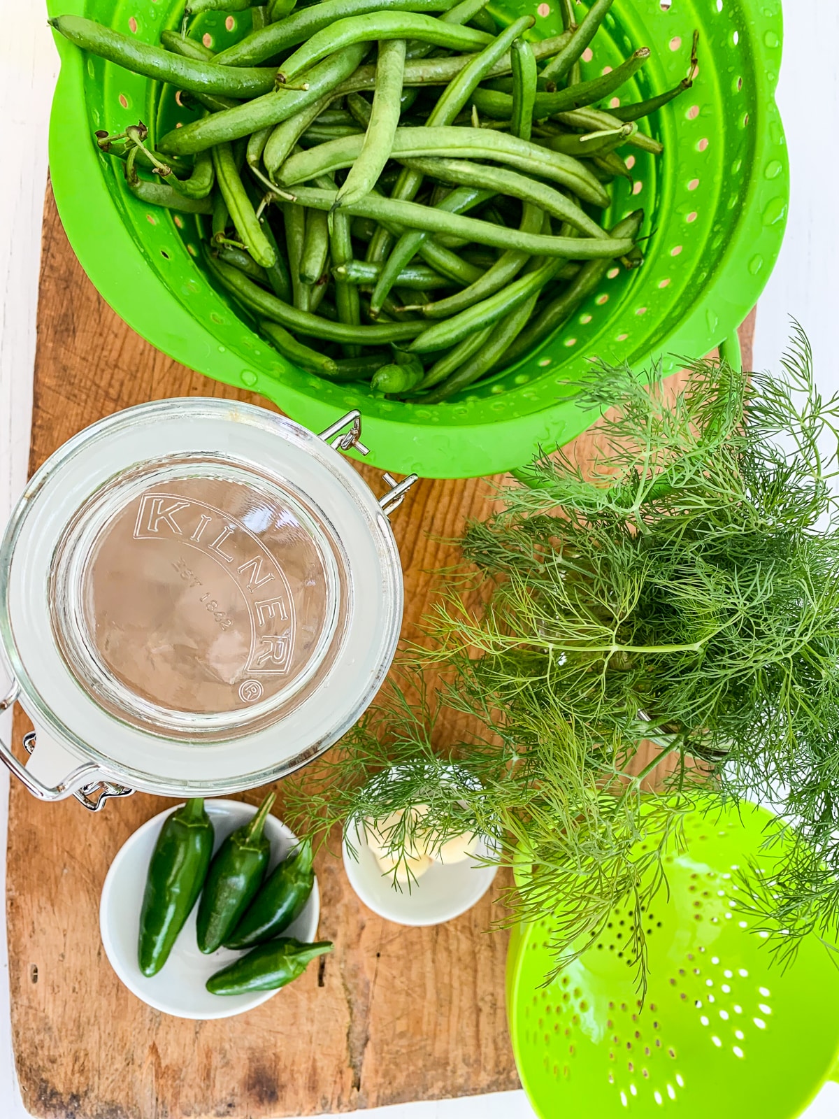 A cutting board with a green colander filled with green beans and ingredients. 