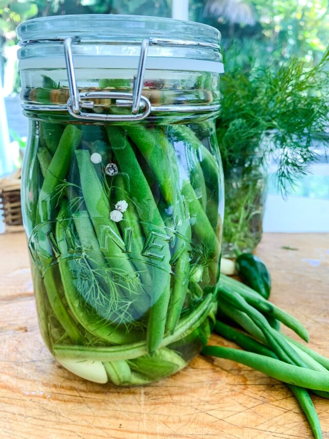 A clear glass clamp jar filled with green bean pickles on a cutting board.