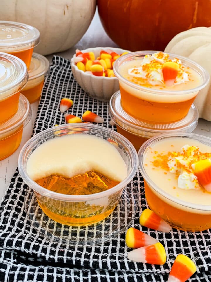 Close up of a candy corn Jello shot that is half sipped and others in plastic cups at a party with candy corn in the background.