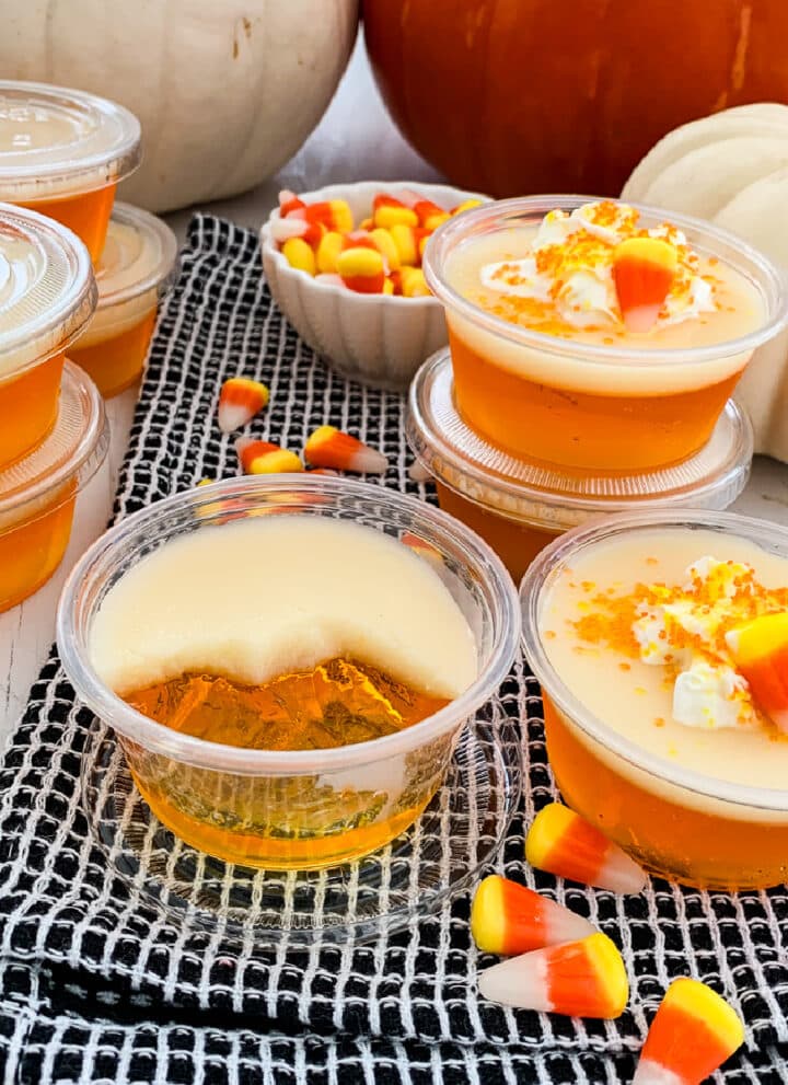 Close up of a candy corn Jello shot that is half sipped and others in plastic cups at a party with candy corn in the background.