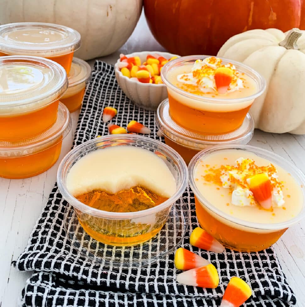 Close up of a candy corn Jello shot that is half sipped and others in plastic cups at a party with candy corn in the background.