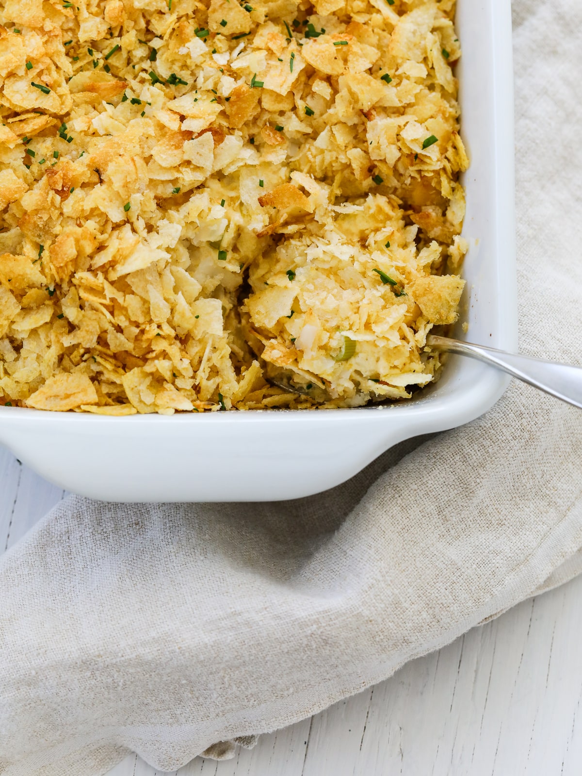 A rectangle white casserole dish with cheesy potato casserole being scooped out with a big serving spoon. 