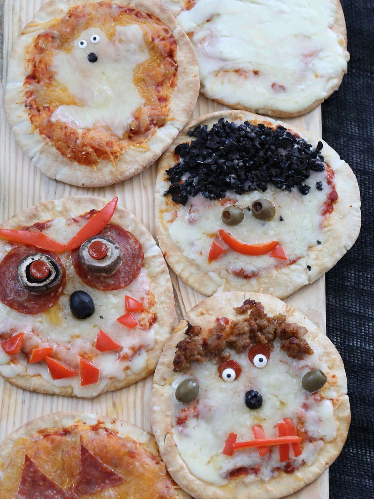 6 different Halloween pizzas baked on a board decorated like jack o lantern, Dracula, mummy, ghost, Frankenstein, and a monster. 